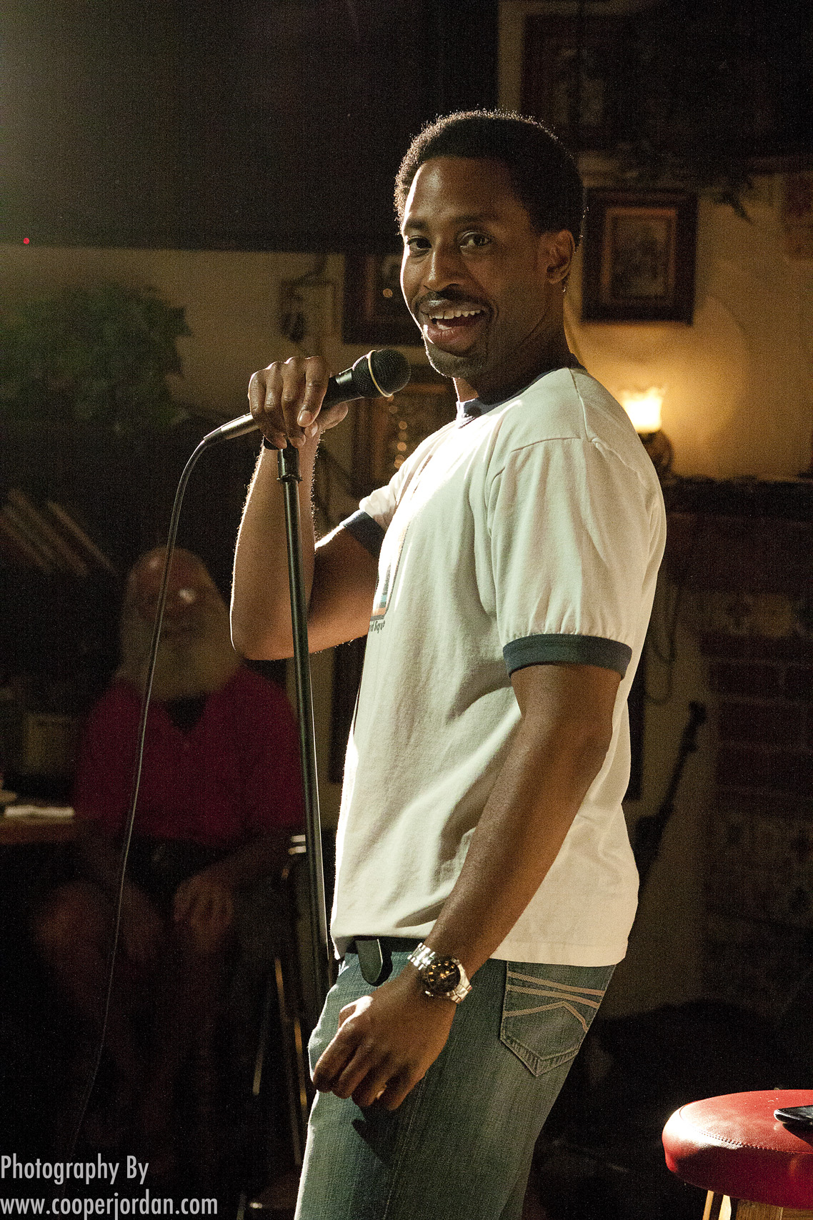 Dwayne Perkins on a comedy club stage in a white t-shirt, grinning at the crowd with a microphone in hand