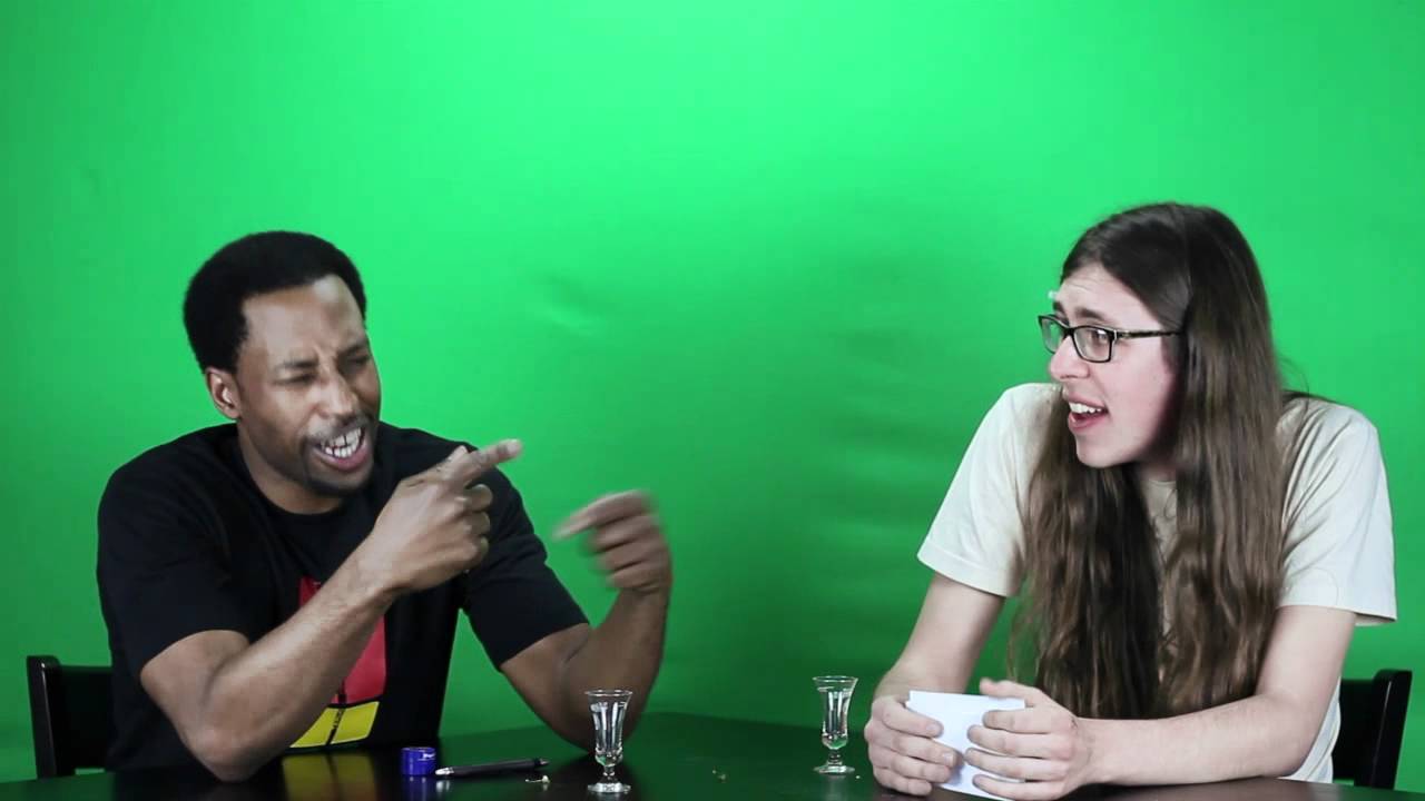 Dwayne Perkins laughing and pointing across a table at a co-host in front of a green screen during a Spicy News sketch